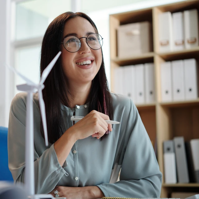 A young woman sits in front of a desk, holding a pen on her right hand and smiling. Next to her, there is a miniature version of a wind fan.